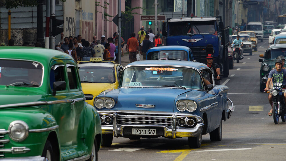 Taxi azul clásico sobre calle con otros autos antiguos y peatones en La Habana.