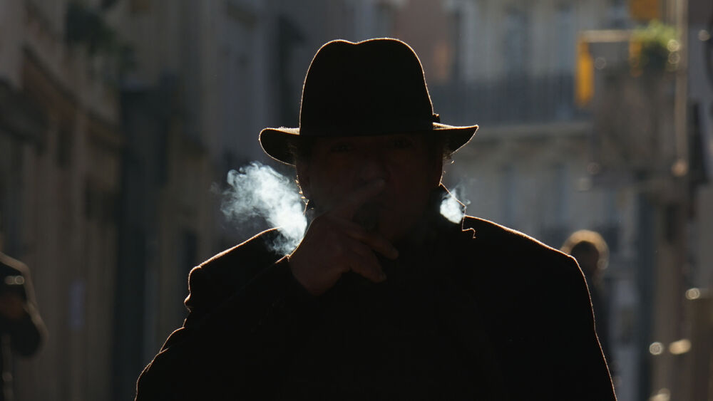 Hombre con sombrero y cigarro emitiendo humo, silueta oscura en calle con luz de fondo.