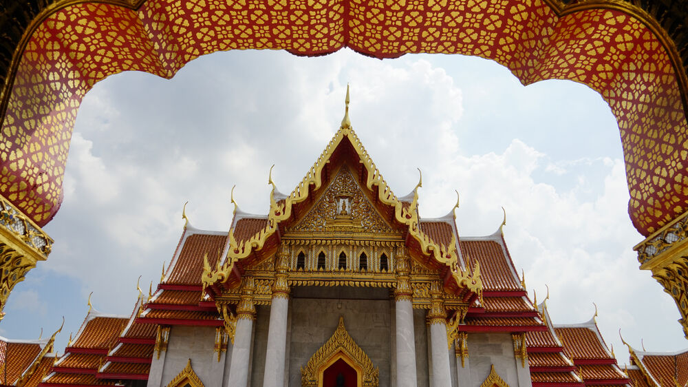 Templo tailandés dorado con techo a dos aguas y adornos florales rojos bajo un cielo nublado.