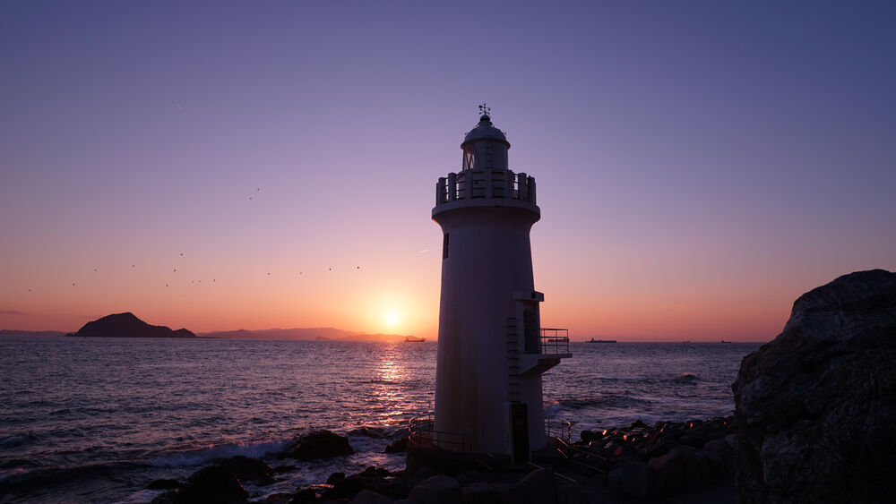 Faro blanco al amanecer sobre el mar, con paisaje costero y cielo púrpura.