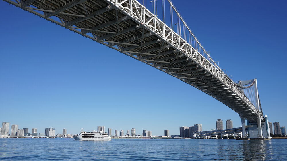 Puente colgante de acero sobre agua azul con skyline de edificios y barco blanco.