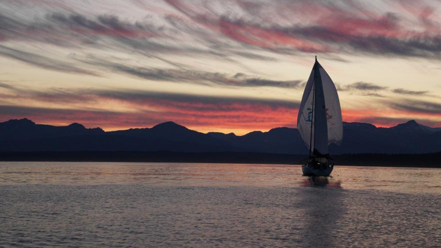 Velero navegando en aguas tranquilas al atardecer, silueta de montañas al fondo, cielo dramático con nubes rosadas y anaranjadas.