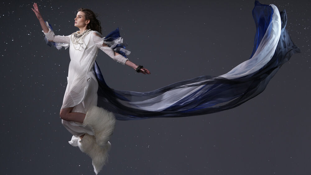 Mujer levitando con vestido blanco plisado, cola de tela azul y blanca, y detalle peludo blanco.
