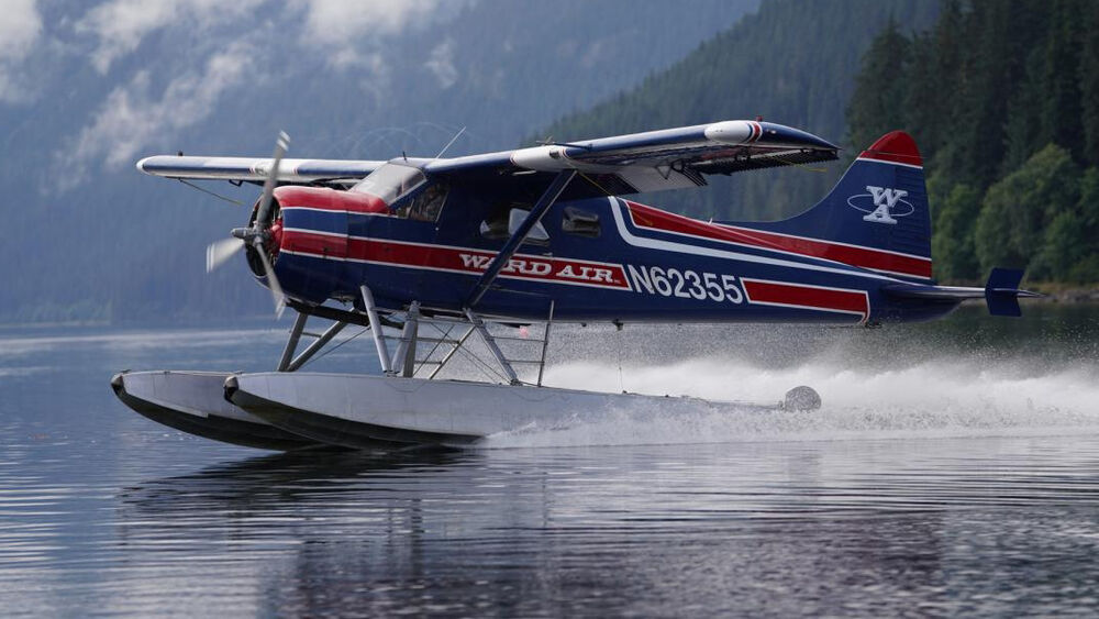 Hidroavión azul y rojo de Ward Air con matrícula N62355 despegando en agua con montañas boscosas de fondo.