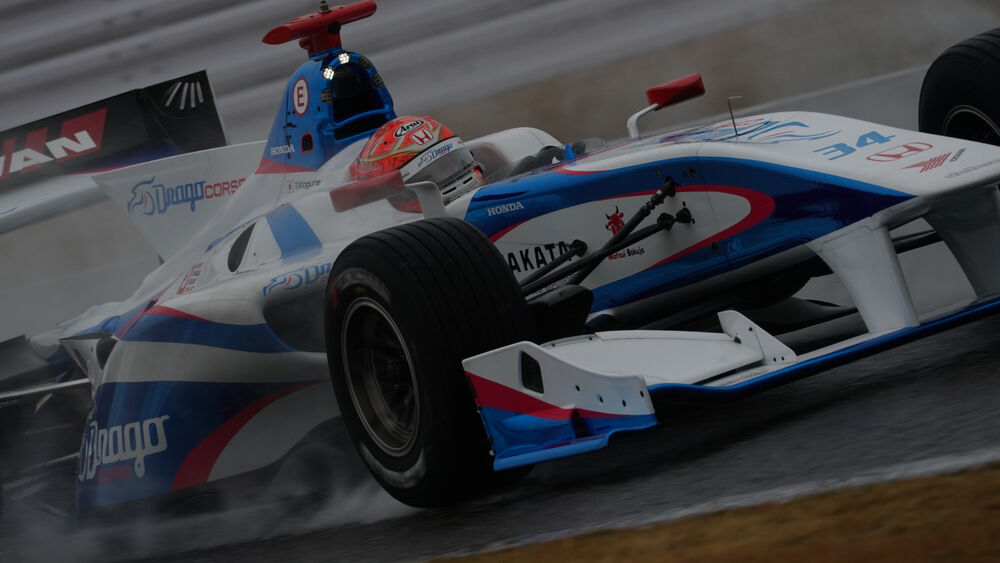 Automóvil de carreras de Fórmula Indy azul y blanco con salpicaduras de agua en pista mojada.