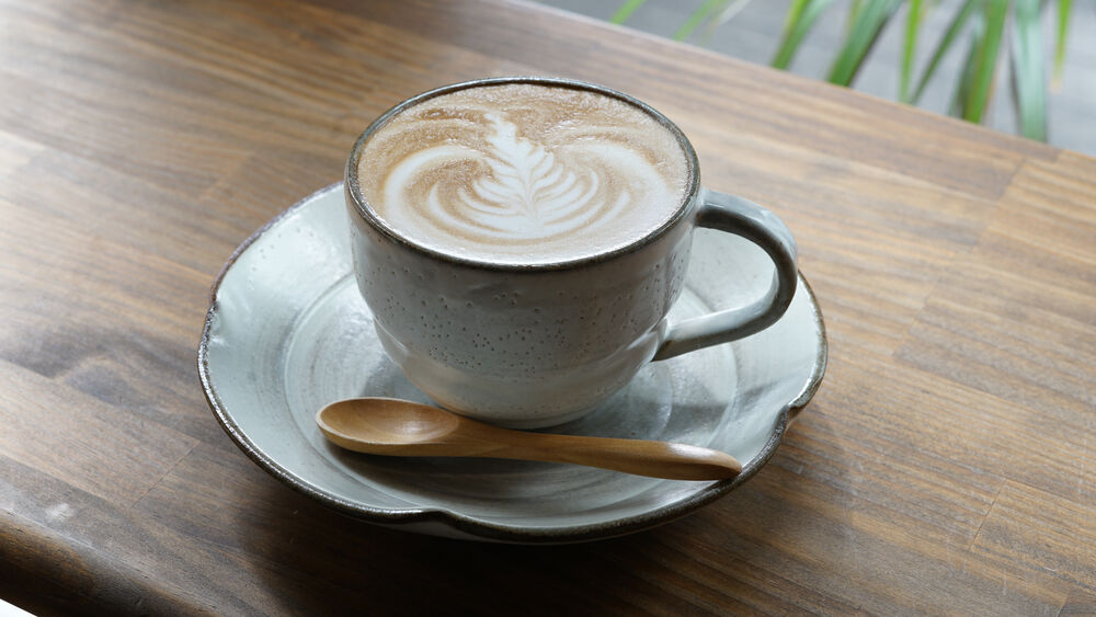 Taza de café latte art en cerámica texturizada sobre plato y cuchara de madera sobre mesa rústica.